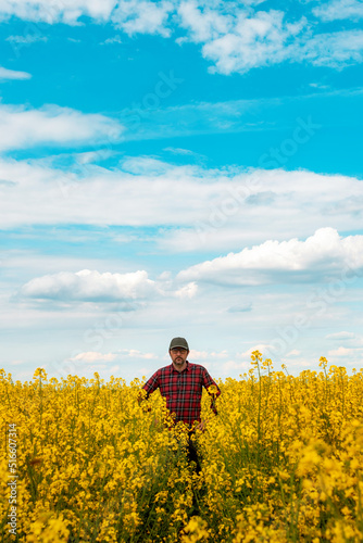 Farm worker wearing red plaid shirt and trucker's hat standing in cultivated rapeseed field in bloom and looking over crops © Bits and Splits