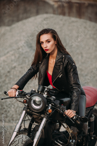  beautiful girl with long dark hair red lips in a black jacket and black pants sits on a black vintage motorcycle in an industrial zone © Катя Іваськевич