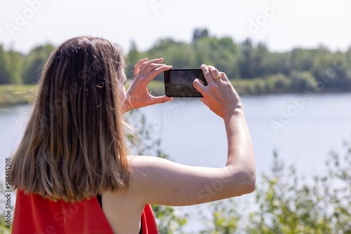 The pretty young caucasian woman in red skirt is taking taking photo by mobile phone in the green forest lakeside at the outside with flare. High quality photo