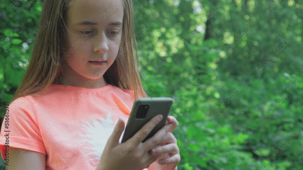 A beautiful teenage girl with long hair scrolls in the phone against the background of tall green trees A beautiful teenage girl with long hair scrolls in the phone against the background of tall green trees
