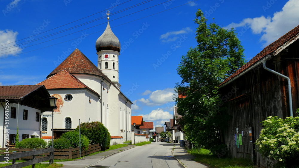 idyllische Dorfstraße in Unterammergau in Bayern mit Kirche, Stall, Baum und Häusern unter blauem Himmel