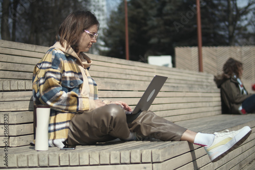 Young adult female is working something on the laptop, while she is sitting on a bench near walkway. Young woman freelancer are working with laptop outdoor in the city
