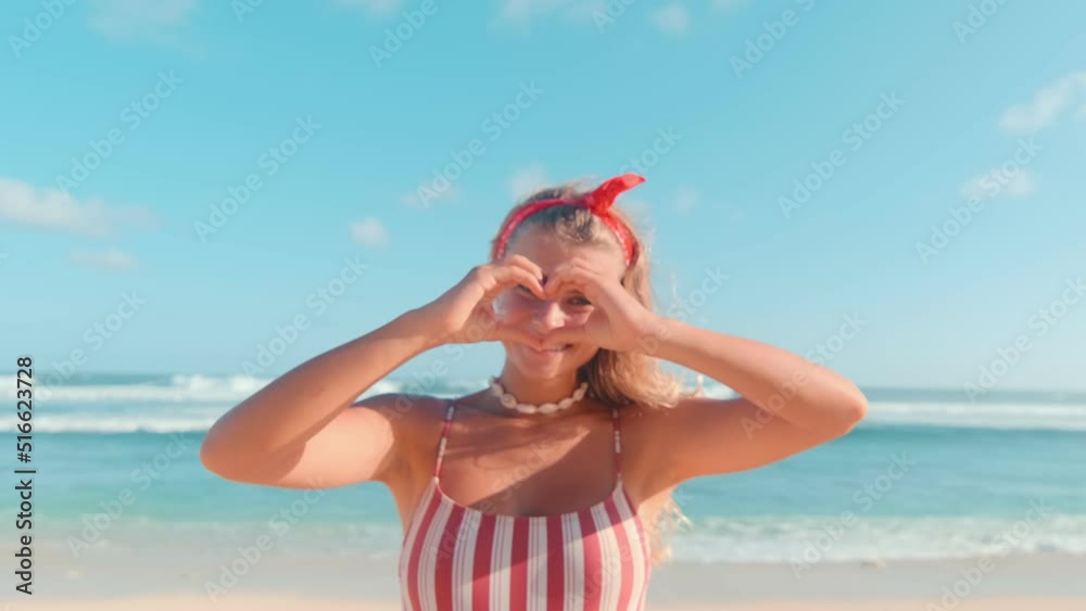 Happy young caucasian woman shows heart shape from fingers sends greetings to family or boyfriend from hot seaside resort dressed in striped swimsuit posing stands on beach of ocean shore Happy young caucasian woman shows heart shape from fingers sends greetings to family or boyfriend from hot seaside resort dressed in striped swimsuit posing stands on beach of ocean shore