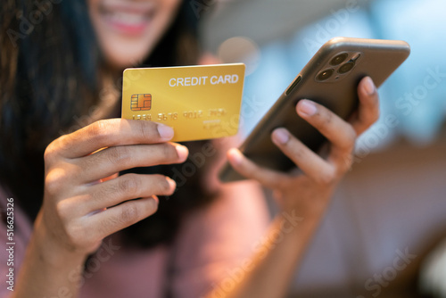 Close-up or closeup of young woman's hand is holding gold credit card and using smartphone with happiness feeling. Female shopper flip her credit card to make purchase online transactions.
