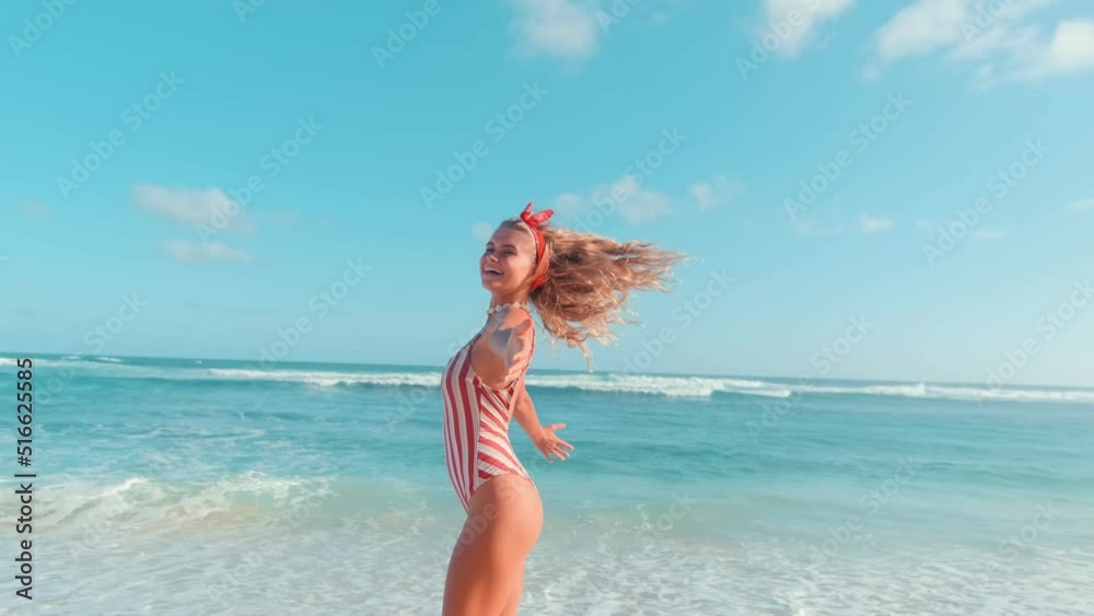 Positive young blonde Caucasian woman student with bow on hair spinning around herself rejoicing at arrival on sunny beach and opportunity to swim or sunbathe dressed in swimsuit enters ocean water Positive young blonde Caucasian woman student with bow on hair spinning around herself rejoicing at arrival on sunny beach and opportunity to swim or sunbathe dressed in swimsuit enters ocean water