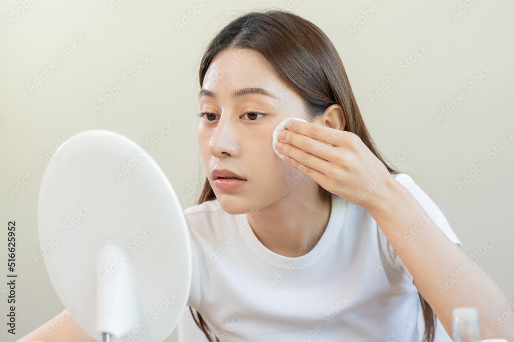Happy beauty, beautiful asian young woman, girl looking in to mirror, holding cotton pad, applying facial wipe on her face, removing makeup before shower in bathroom, skin care on white background. Happy beauty, beautiful asian young woman, girl looking in to mirror, holding cotton pad, applying facial wipe on her face, removing makeup before shower in bathroom, skin care on white background.