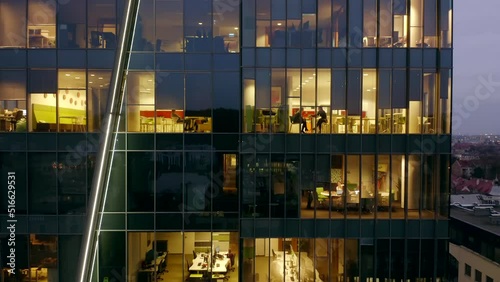 Office building at night with people working inside night skyscraper aerial view. Working evening in the office building of a glass business center with numerous offices.