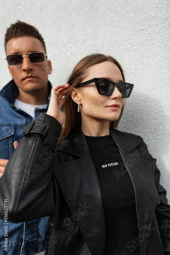 Stylish beautiful young woman model with vintage fashion sunglasses in a fashionable black coat and T-shirt and a young handsome man in fashionable denim clothes on the street. Fashionable couple