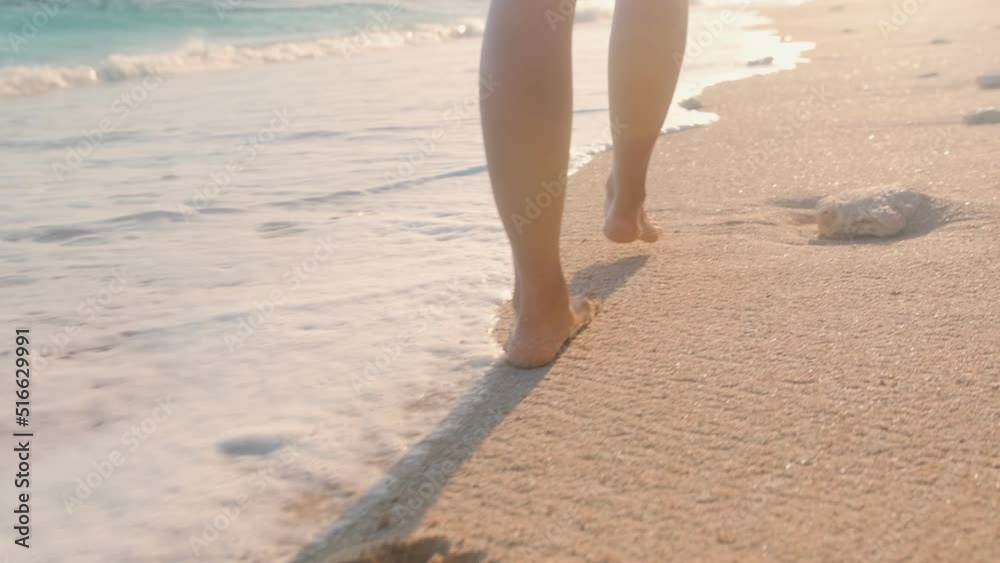 Close up beautiful bare feet woman walking on sea sand at high tide passing next to waves of ocean leaving footprints on ground be on sunny beach. Summer holidays, recreation, tourism
