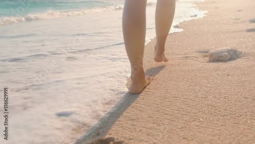 Close up beautiful bare feet woman walking on sea sand at high tide passing next to waves of ocean leaving footprints on ground be on sunny beach. Summer holidays, recreation, tourism photo