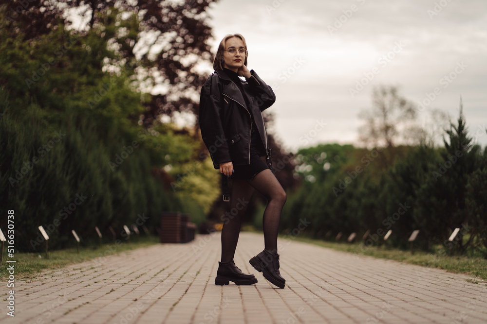 Cute happy girl with glasses in a black jacket and dress walks in the park alone in autumn. Pretty young girl with blond hair in black walks alone in the park and poses on the empty road. Cute happy girl with glasses in a black jacket and dress walks in the park alone in autumn. Pretty young girl with blond hair in black walks alone in the park and poses on the empty road.