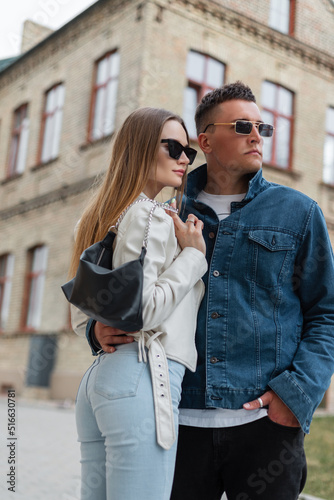 Fashion street beautiful young stylish couple guy and woman hipster with vintage sunglasses in fashionable denim clothes with a purse walking together in the city