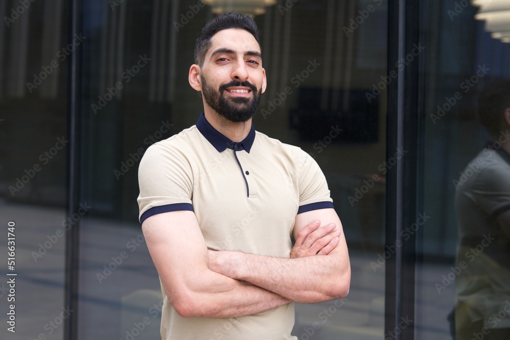 Portrait of happy positive confident successful Arab African Muslim ethnic guy, young bearded businessman, man with black beard standing outdoors smiling looking at camera with his hands crossed - obrazy, fototapety, plakaty 