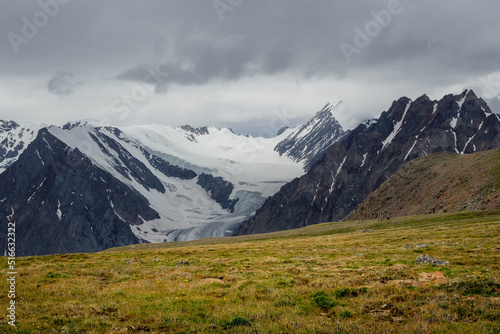 Summer view from sunlitgreen grass hill to high snowy mountain range with sharp tops and glaciers under cloudy sky. Colorful landscape with large snow mountains at changeable weather.