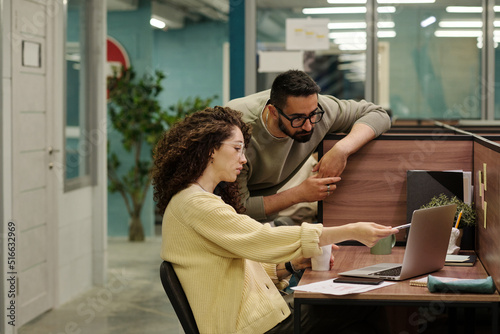 Young confident businesswoman pointing at data on laptop screen while making presentation or slide show of points of new project to colleague