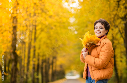 Cheerful young girl with short brown hair wearing autumn yellow jacket  walking at the park. Autumn walk. Woman portrait. Girl is playing with leaf and smiling. Woman enjoying fall nature.