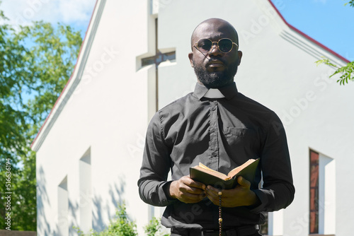 Young black man in shirt with clerical collar standing with open Bible in front of camera while preaching in front of church building