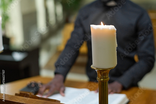 White burning candle on background of priest reading verses from Holy Bible while standing by pulpit and telling sermon during church service photo
