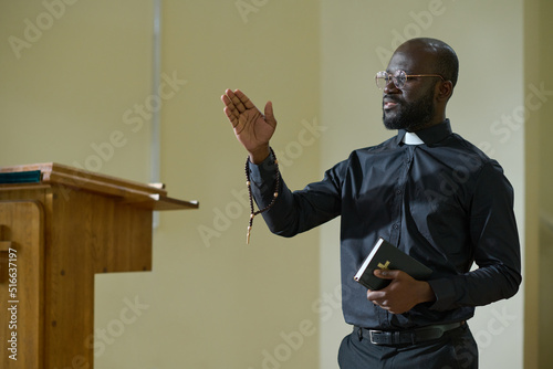 Confident African American man in pastor apparel with clerical collar preaching during church service in front of parishioners photo