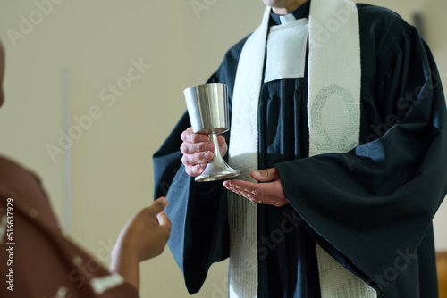 Clergyman of Catholic church holding cup with wine for oblation rite while standing in front of African American female parishioner
