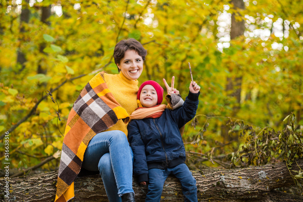 Mother and her little son are sitting on a log in the autumn park, posing, smiling and looking at the camera Mother and her little son are sitting on a log in the autumn park, posing, smiling and looking at the camera