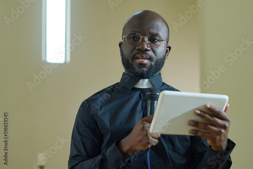 Young confident black man with microphone and digital tablet making speech for parishioners of evengelical church during sermon photo