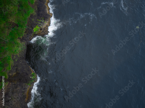 Fototapeta Naklejka Na Ścianę i Meble -  Top view of a calm dark ocean surface with white light waves near a hilly green shore. The beauty of pristine nature. Deserted place. Geology, weather, environmental protection.