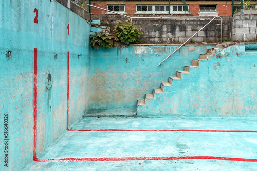 Abandoned public accessible swimming pool, focus on corner with stairs. The pool has a bright blue colour with red stripes.
