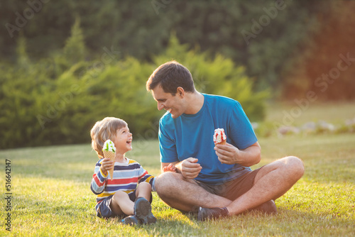 Laughing young father and little son haveing fun together and eating ice cream, sitting on grass in green park on summer day. Happy family concept