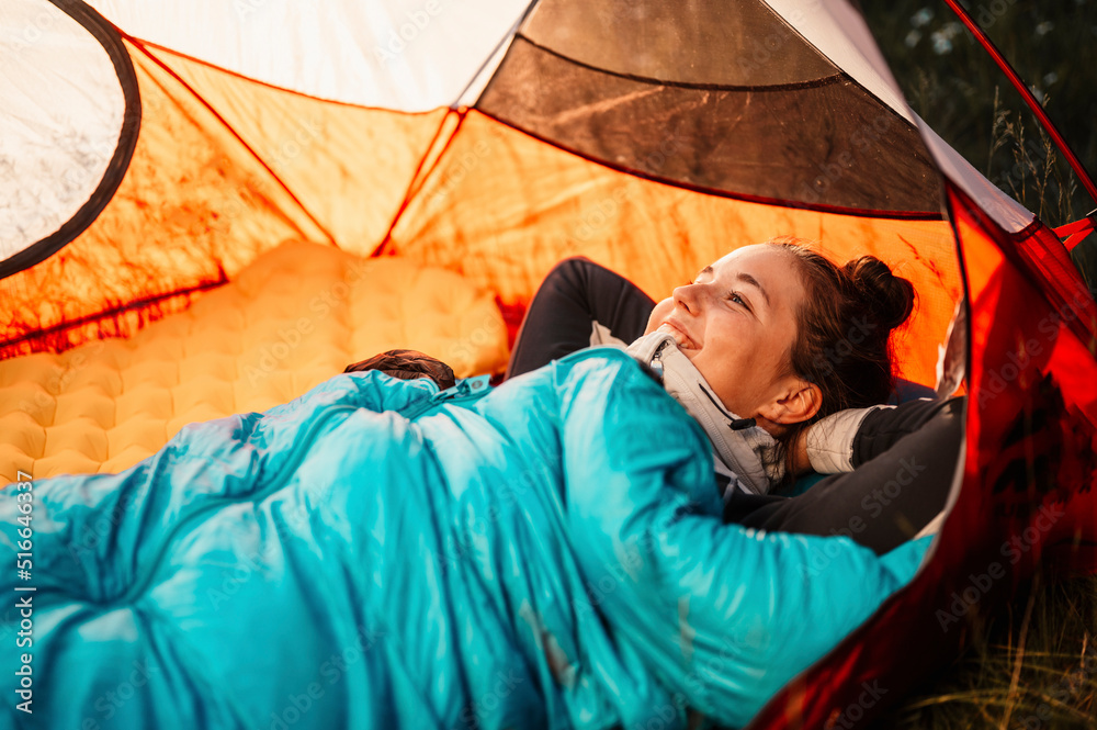 Woman relaxing and lie in a sleeping bag in the tent. Sunset camping in forest. Mountains
