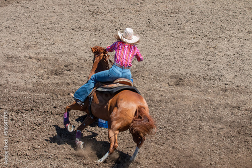 A Rodeo barrel racer is rounding a blue barrel. The cowgirl has a white hat, red shirt and blue pants. The horse has a reddish coat. The area is dirt. photo