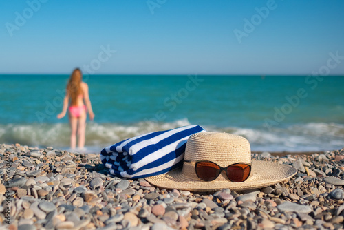 Hat, a towel and sunglasses. Tall woman with a long hair from a back is on a beach. Mediterranean sea with waves on the background. Blue and turquoise water. Vacation summer vibe. Slow motion video. 