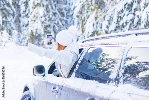teenage girl in white sweater, vest and white knitted hat in car window in snowy forest take selfie photo on mobile phone, concept winter local travel during Christmas or New Year holidays vacation