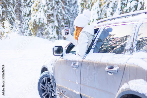 teenage girl in white sweater  vest and white knitted hat in car window in snowy forest take selfie photo on mobile phone  concept winter local travel during Christmas or New Year holidays vacation