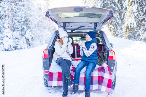 two children friends boy and girl teenagers traveling and having fun in trunk car in snow winter forest, drinking tea from thermos, talking and get ready to celebrate Christmas or New year together