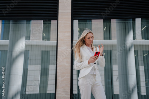 Pretty young lawyer in white suite standing outdoors against contemporary building with large windows making video call. Italian confident entrepreneur arguing with partner, agent convinces client.