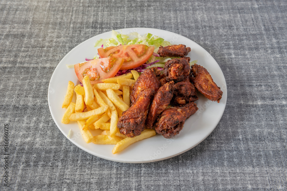 Fried chicken wings with salad and tomato and french fries served in typical kebab restaurant with white plate and gray table Fried chicken wings with salad and tomato and french fries served in typical kebab restaurant with white plate and gray table