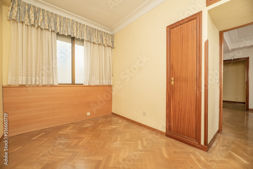 Empty bedroom with herringbone oak floor, light yellow painted walls and built-in wardrobe with wooden door and curtained window