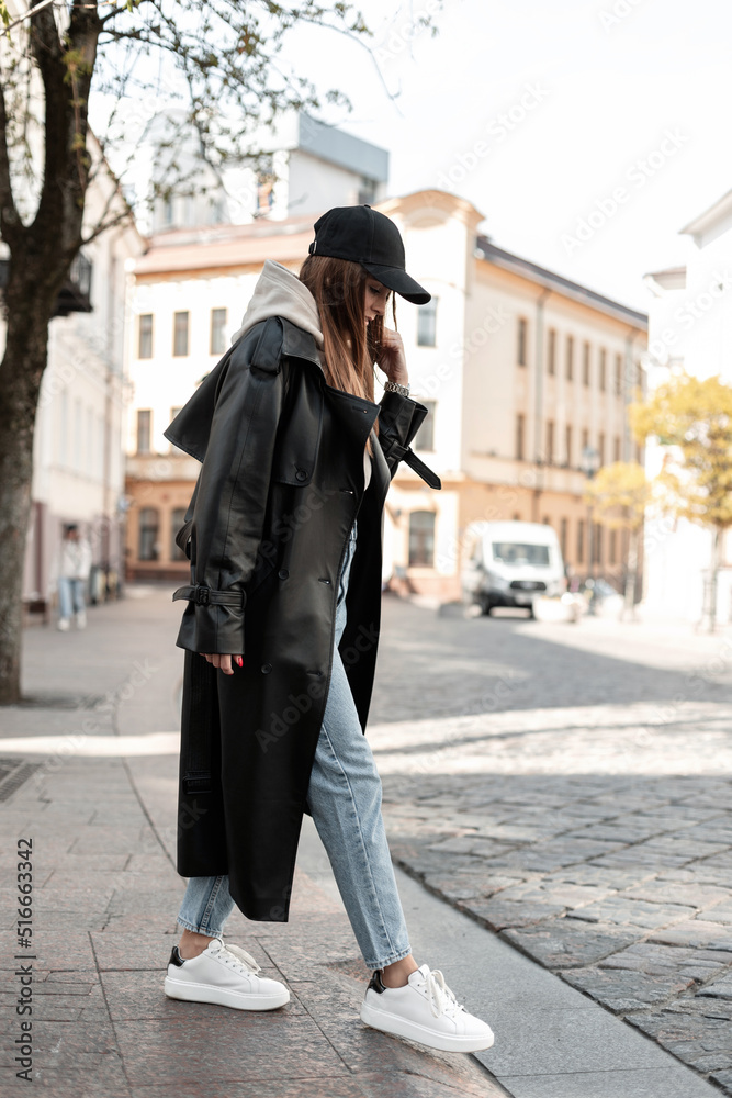 Fashion beautiful young woman model hipster in fashionable black casual clothes with a cap in a long leather coat with jeans and white shoes walking in the city. Cool female urban outfit look