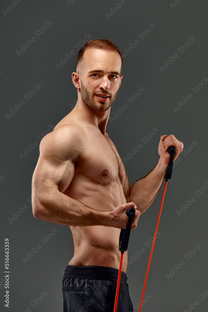 A topless sportsman performs fitness exercises with rubber band in a studio on gray background. Body-building. Athlete. A topless sportsman performs fitness exercises with rubber band in a studio on gray background. Body-building. Athlete.