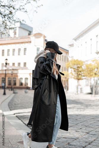 Fashionable beautiful street hipster woman in stylish urban casual clothes with a fashion black long leather coat and cap walking in the city and wearing a hood