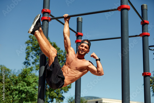 Athletic young man hanging from the bars at the calisthenics gym outdoors smiling