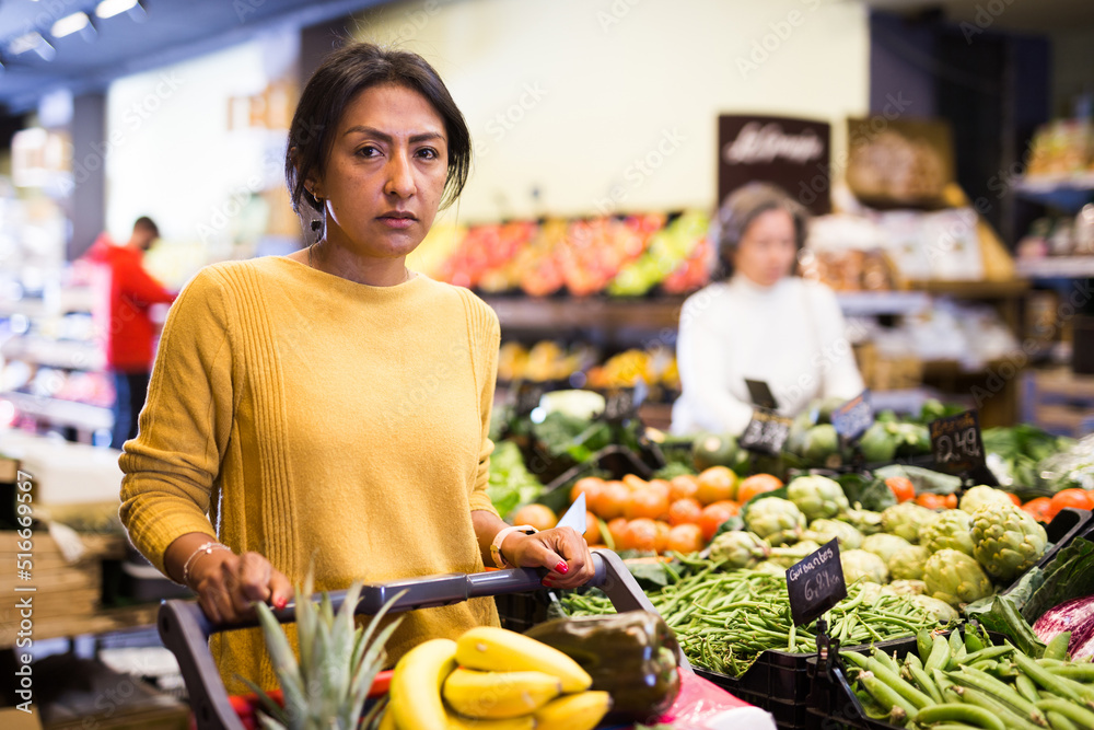 Portrait of thoughtful Hispanic woman walking with shopping trolley among shelves with products in grocery shop
