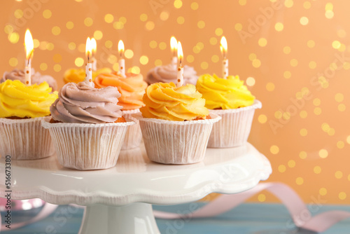 White stand with tasty birthday cupcakes on light blue wooden table against blurred lights, closeup