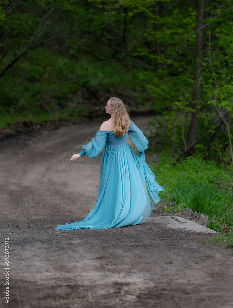 One blonde haired woman wearing long flowing blue gown in green meadow. Magical, fairytale concept.	