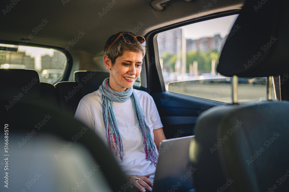 Fototapeta premium one mature woman caucasian female sitting on the back seat of the car working on laptop computer in summer day with short gray hair modern manager or business owner on road wearing casual copy space