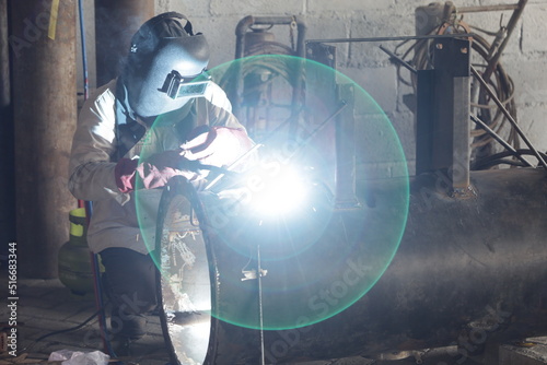 A young man welder in brown uniform  welding mask and welders leathers  weld metal with a arc welding machine at the construction site  blue sparks fly to the sides