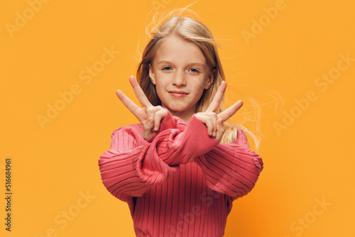 a cute, happy school-age girl stands on a yellow background in a pink sweater and shows a victory sign with her fingers smiling broadly