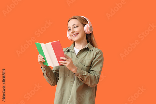 Mature woman with headphones reading book on orange background. Concept of studying Italian language photo
