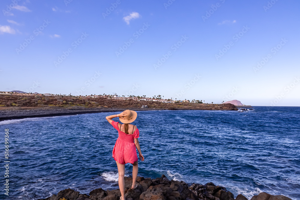 Woman with summer hat and red dress enjoying scenic view on black stone pebble beach Playa Colmenares near Amarilla, Golf del Sur, Tenerife, Canary Islands, Spain, Europe. Waves from Atlantic Ocean